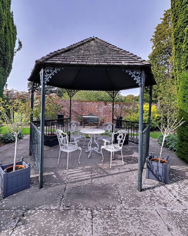 The slate-roofed wrought-iron gazebo on its paved terrace, with white garden chairs inside and the walled garden behind
