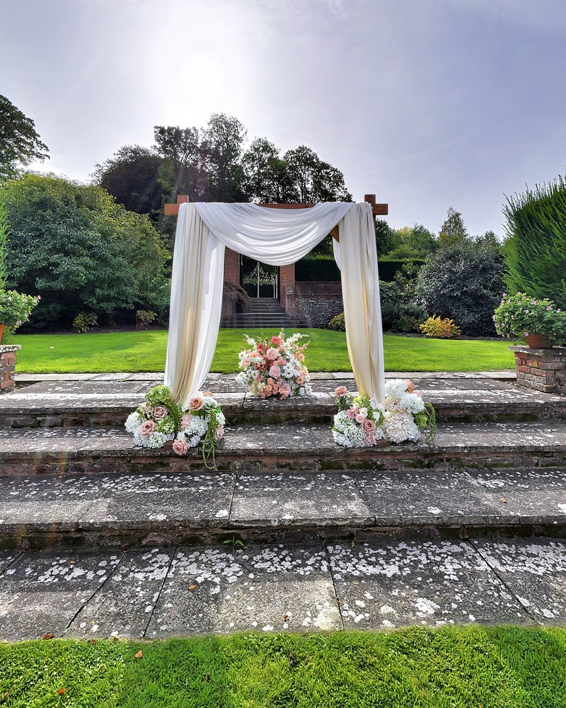 A draped ceremony arch dressed with blush roses at the top of wide stone steps, the lawn and walled garden beyond