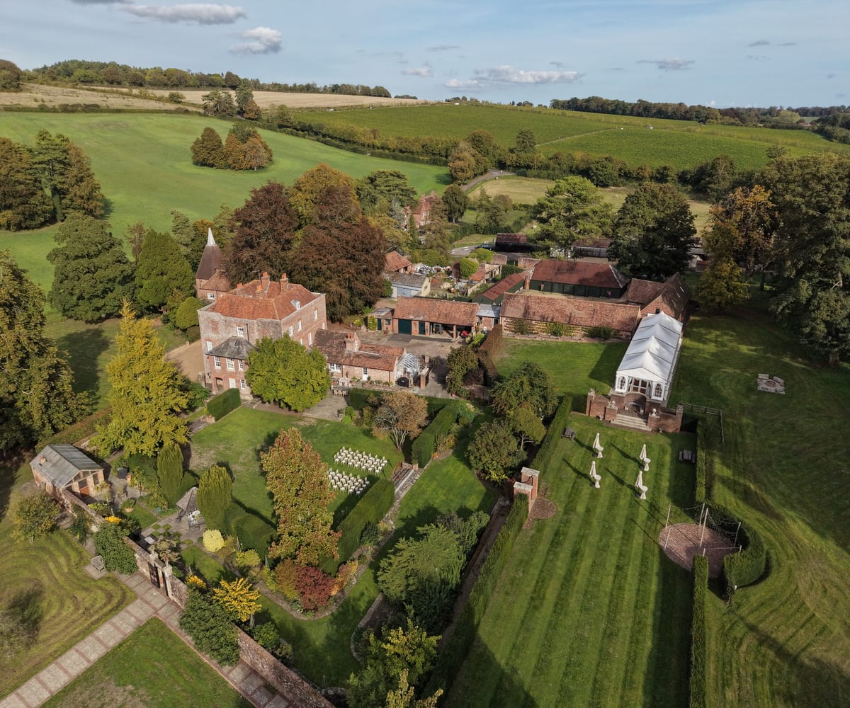 Aerial view of Swarling Manor: the red-brick house and barns, the white marquee, striped lawns and walled gardens, with rolling Kent farmland beyond
