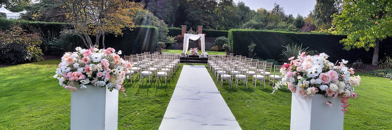 A white aisle runner between rows of chiavari chairs, framed by two pedestals of blush and white roses, leading to the draped arch on the Steps