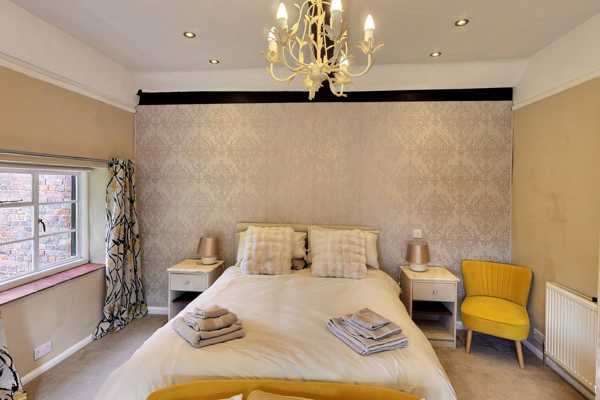 A double bedroom at Coach Cottage with a chandelier, damask feature wall and a mustard armchair