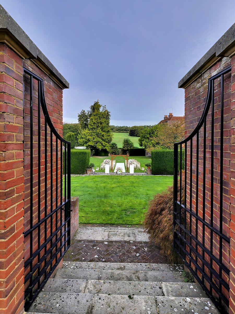 Open wrought-iron gates between red-brick piers, looking down stone steps to the ceremony lawn