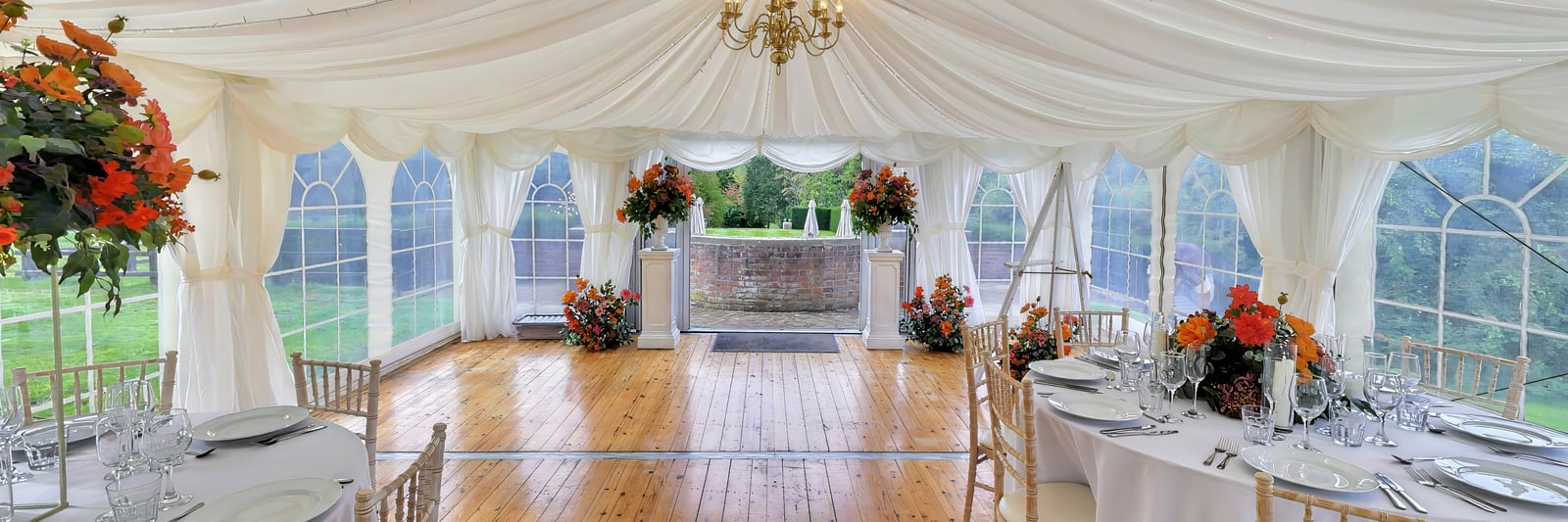 The marquee laid for the wedding breakfast: white linen, chiavari chairs and orange florals beneath a draped ceiling and chandelier