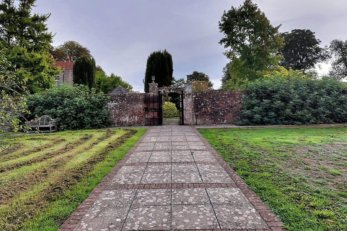 A paved path running straight between fruit trees and long grass to a gateway in the old garden wall